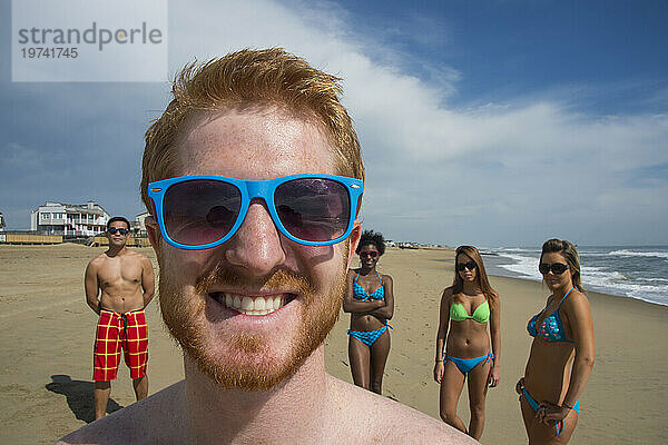 Gruppe von Freunden in Badebekleidung am Virginia Beach  mit einem jungen Mann im Vordergrund mit einem breiten Lächeln und einer blauen Sonnenbrille  First Landing State Park  Virginia  USA; Virginia Beach  Virginia  Vereinigte Staaten von Amerika