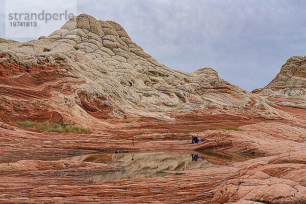 Malerischer Blick auf Navajo-Sandsteinfelsformationen  die als Brain Rocks bezeichnet werden  mit einem auf den Felsen liegenden Fotografen und einer Kamera auf einem Stativ  die sich in einem Wasserbecken unter bewölktem Himmel in der wundersamen Gegend von White Pocket mit seinen fremdartigen Landschaften aus erstaunlichen Linien spiegelt. Konturen und Formen; Arizona  Vereinigte Staaten von Amerika