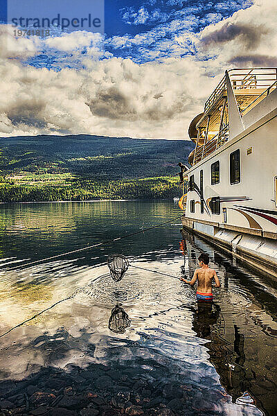 Eine Familie genießt einen Hausbooturlaub  während sie am Ufer des Shuswap Lake parkt  mit einem Mann  der vom Deck des Hausboots aus fischt  und einem Jungen  der im Badeanzug im Wasser steht und ein Fischernetz in der Hand hält; Shuswap Lake  British Columbia  Kanada