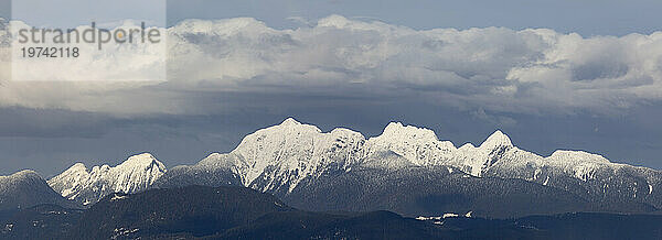 Dramatische Aussicht auf einen Kamm schneebedeckter Berggipfel der Coast Mountains  gesehen von Surrey  BC  unter einem wolkenverhangenen blauen Himmel; Surrey  British Columbia  Kanada