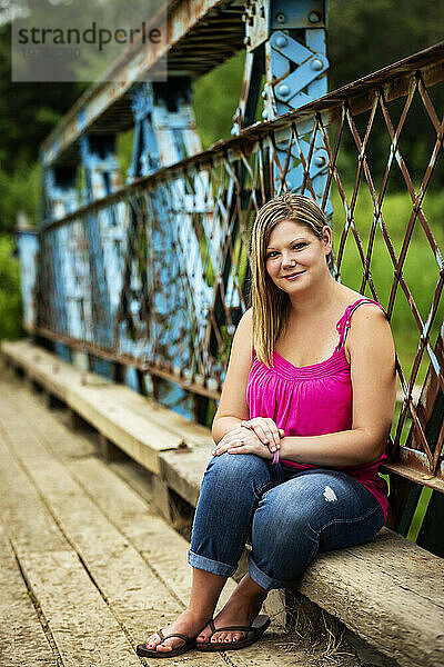 Nahaufnahmeporträt einer Frau  die während eines Naturspaziergangs in einem Park auf einer Holzbank entlang einer Bockbrücke sitzt und für die Kamera posiert; Edmonton  Alberta  Kanada