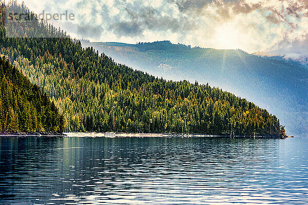 Ein Hausboot  das am Ufer des wunderschönen Shuswap-Sees geparkt ist  mit Sonnenstrahlen  die während der Herbstsaison dramatische Beleuchtung und Reflexionen über den Bergen und dem Wasser erzeugen; Shuswap Lake  British Columbia  Kanada