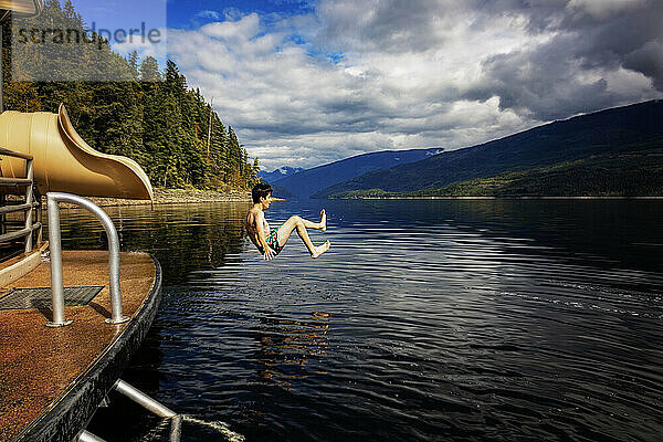 Junge springt an einem Herbsttag auf dem Shuswap Lake aus dem Ende einer Wasserrutsche auf einem Hausboot; British Columbia  Kanada