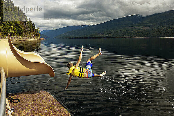 Kleiner Junge springt an einem Herbsttag auf dem Shuswap Lake aus dem Ende einer Wasserrutsche auf einem Hausboot; British Columbia  Kanada