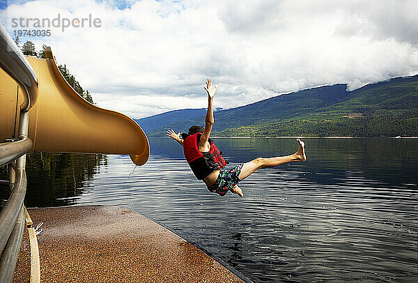 Kleiner Junge springt an einem Herbsttag auf dem Shuswap Lake aus dem Ende einer Wasserrutsche auf einem Hausboot; British Columbia  Kanada