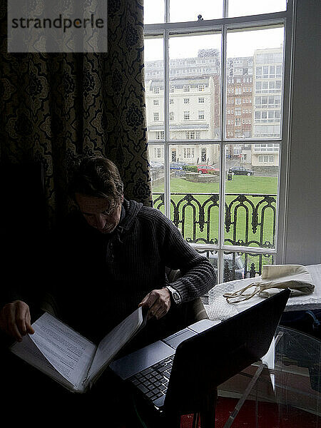 Mann sitzt an einem regnerischen Tag mit Laptop am Fenster und liest Dokumente in einem Hotelzimmer in Brighton; Brighton  East Sussex  England  Vereinigtes Königreich