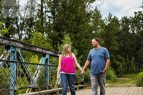 Porträt eines Paares  das auf einer hölzernen Bockbrücke läuft und sich bei einem Naturspaziergang in einem Park Händchen haltend anschaut; Edmonton  Alberta  Kanada