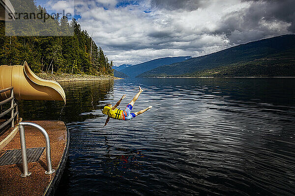 Kleiner Junge springt an einem Herbsttag auf dem Shuswap Lake aus dem Ende einer Wasserrutsche auf einem Hausboot; British Columbia  Kanada