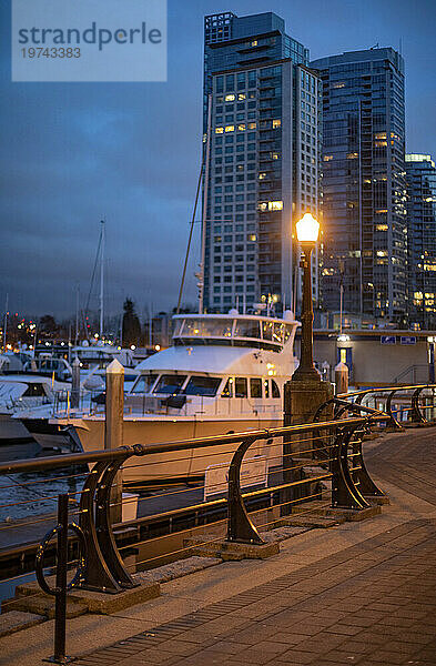 Nahaufnahme von im Jachthafen vertäuten Booten und der von einem Laternenpfahl beleuchteten Promenade mit Hochhäusern im Hintergrund an der Uferpromenade des Hafens von Vancouver in der Dämmerung; Vancouver  British Columbia  Kanada