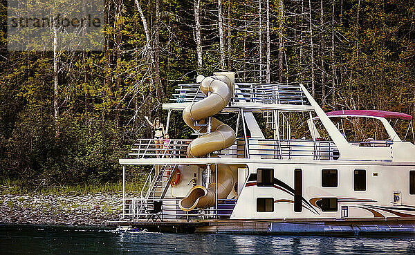 Eine Familie  die einen Hausbooturlaub genießt  während sie am Ufer des Shuswap Lake parkt  mit Mutter und Tochter  die vom Deck am Heck des Bootes winken  und einem Jungen im Teenageralter  der im Hintergrund an Deck sitzt; Shuswap Lake  British Columbia  Kanada