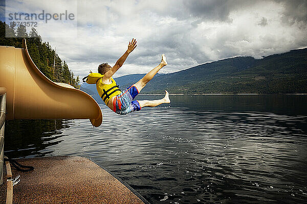 Kleiner Junge springt an einem Herbsttag auf dem Shuswap Lake aus dem Ende einer Wasserrutsche auf einem Hausboot; British Columbia  Kanada