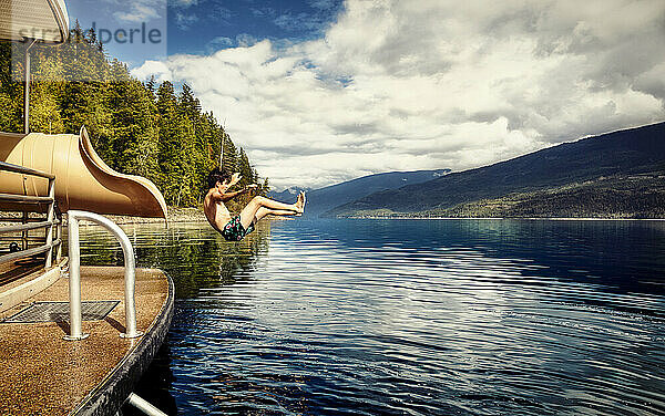 Junge springt an einem Herbsttag auf dem Shuswap Lake aus dem Ende einer Wasserrutsche auf einem Hausboot; British Columbia  Kanada