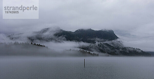 Atemberaubender Blick von einem Spaziergang entlang des Deichs am Pitt Lake in Maple Ridge auf einen Berggipfel  der an einem grauen Tag in nebligen Nebel gehüllt ist; Maple Ridge  British Columbia  Kanada