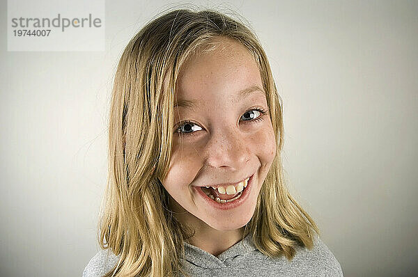 Porträt eines jungen Mädchens mit blonden Haaren und blauen Augen  das vor einem grauen Hintergrund ein breites Lächeln in die Kamera wirft; Studio