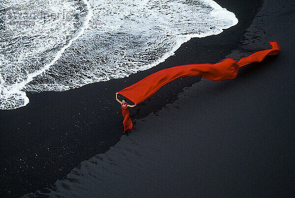 Frau in fließendem Rot an einem schwarzen Sandstrand am Rande der Brandung in Island; Island