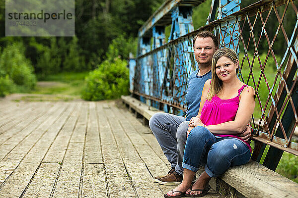 Porträt eines Paares  das bei einem Naturspaziergang in einem Park zusammen auf einer hölzernen Bockbrücke sitzt und in die Kamera schaut; Edmonton  Alberta  Kanada