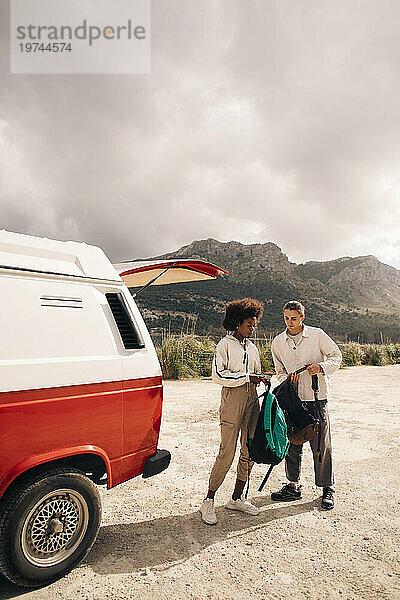 Männliche und weibliche Freunde halten Rucksäcke in der Hand  während sie in der Nähe eines Lieferwagens vor den Bergen stehen