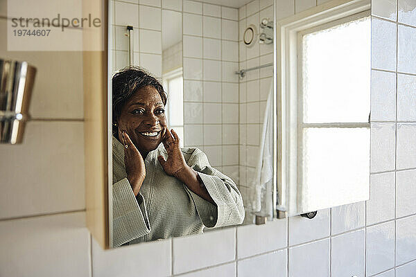 Glückliche reife Frau berührt ihr Gesicht  während sie sich zu Hause im Badezimmerspiegel betrachtet