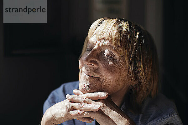 Lächelnde blonde Seniorin mit den Händen am Kinn entspannt sich zu Hause im Sonnenlicht