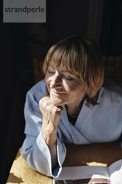 Blonde ältere Frau mit der Hand am Kinn entspannt sich zu Hause im Sonnenlicht