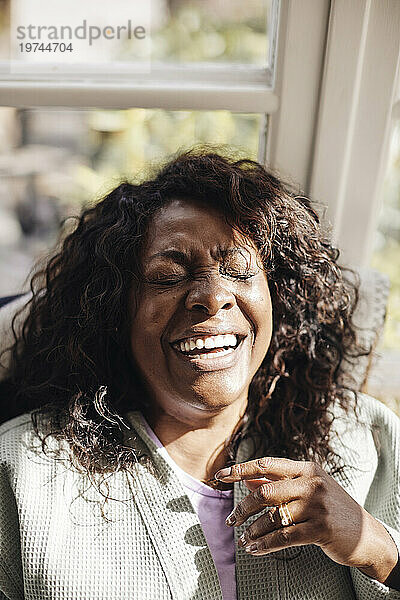 Reife Frau mit geschlossenen Augen lacht zu Hause im Sonnenlicht