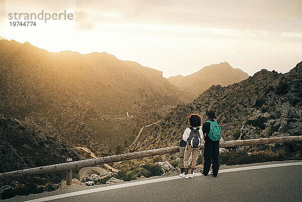 Mann und Frau blicken auf Berge  während sie bei Sonnenuntergang in der Nähe eines Geländers auf der Straße stehen