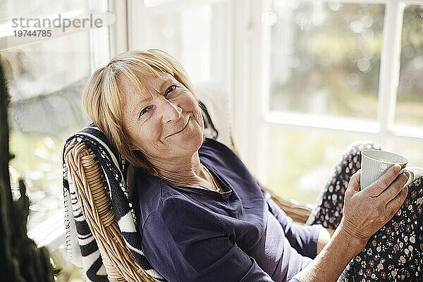 Porträt einer lächelnden blonden Seniorin mit Kaffeetasse  die zu Hause auf einem Stuhl sitzt