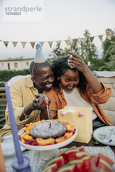 Fröhlicher junger Mann sitzt mit seiner Freundin zusammen und schneidet Kuchen an  während er im Hinterhof seinen Geburtstag feiert