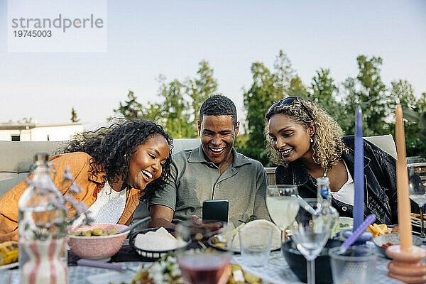 Lächelnder Mann  der während der Dinnerparty im Hinterhof sein Smartphone mit Freundinnen teilt