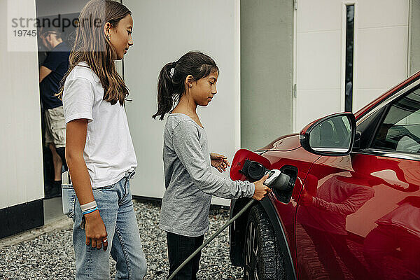 Mädchen steht neben Schwester und lädt Elektroauto in der Nähe des Hauses auf