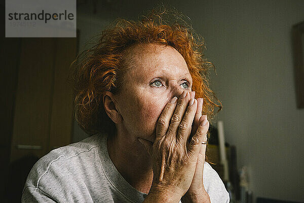 Nachdenkliche  traurige ältere Frau mit Händen  die den Mund bedecken  sitzt zu Hause