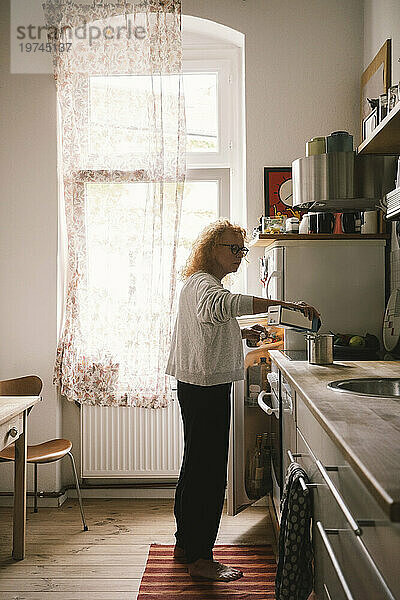 Seitenansicht einer älteren Frau  die Milch in einen Behälter gießt  während sie zu Hause in der Küche steht