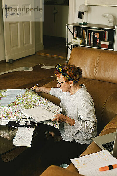 Seitenansicht einer älteren Frau  die auf der Karte eine Reise plant  während sie zu Hause im Wohnzimmer sitzt
