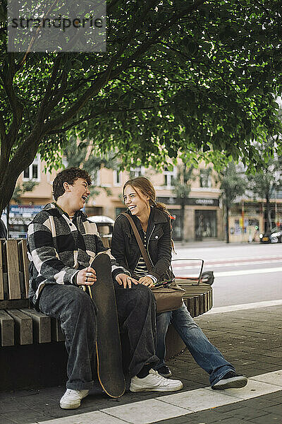 Glückliche männliche und weibliche Teenager-Freunde lachen  während sie auf einer Bank unter einem Baum sitzen