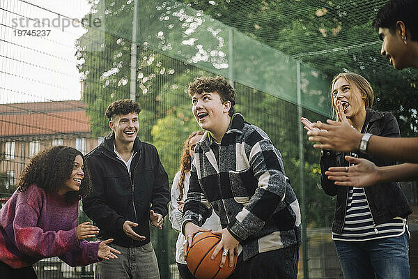 Fröhliche Freunde jubeln einem Teenager zu  der sich auf den Schuss vorbereitet  während er auf dem Platz Basketball spielt