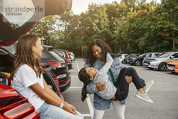 Mädchen sitzt im Kofferraum und schaut der Mutter zu  die sich mit ihrer Tochter in der Nähe des Autos amüsiert