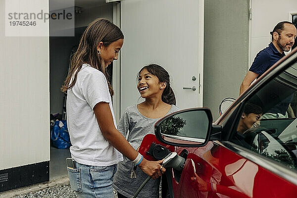 Lächelndes Mädchen schaut Schwester zu  die in der Nähe des Hauses ein Elektroauto auflädt