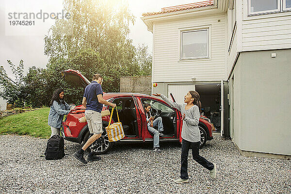 Mädchen spielt mit Ball in der Nähe einer Familie  die am Haus Sachen in ein Elektroauto lädt