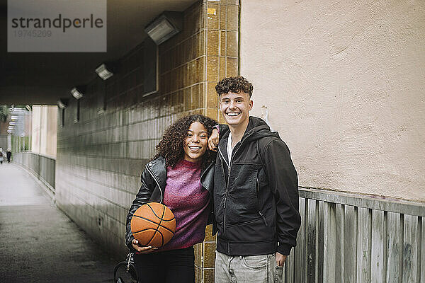 Porträt eines lächelnden Mädchens  das Basketball hält  während es mit einem männlichen Freund in der Nähe einer Unterführung steht