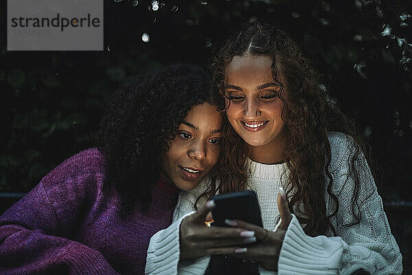 Lächelndes Mädchen teilt Smartphone mit neugieriger Freundin