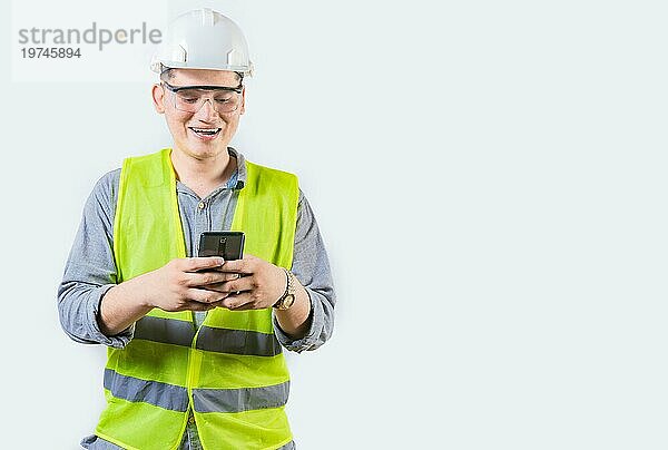 Hübscher Ingenieur hält Telefon isoliert. Fröhlich Bauingenieur texting Telefon auf isolierten Hintergrund. Junger Ingenieur in Weste und Helm mit Mobiltelefon