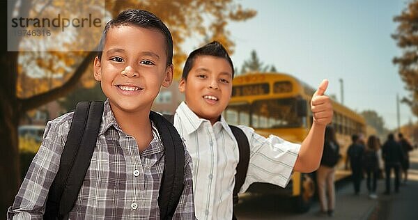 Zwei glückliche hispanische Jungen mit Rucksäcken zeigen auf dem Campus in der Nähe eines Schulbusses die Daumen nach oben