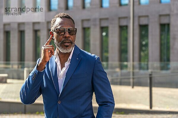 Cooler reifer afrikanischer Geschäftsmann mit Sonnenbrille und Anzug  der über Kopfhörer mit dem Handy im Freien spricht
