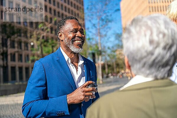 Afrikanischer reifer Geschäftsmann im Gespräch mit Kollegen  der entspannt und lächelnd Kaffee zum Mitnehmen im Freien trinkt