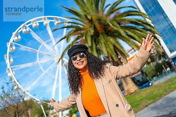 Glückliche modische lateinische Frau neben einem Riesenrad an einem sonnigen Wintertag in der Stadt