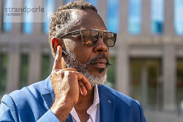 Cooler afrikanischer reifer Geschäftsmann  der die Kopfhörer einstellt  um ein Gespräch mit dem Handy im Freien zu hören