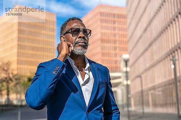 Reifer afrikanischer Geschäftsmann mit Sonnenbrille  der den Kopfhörer einstellt  um im Freien ein Telefongespräch zu führen