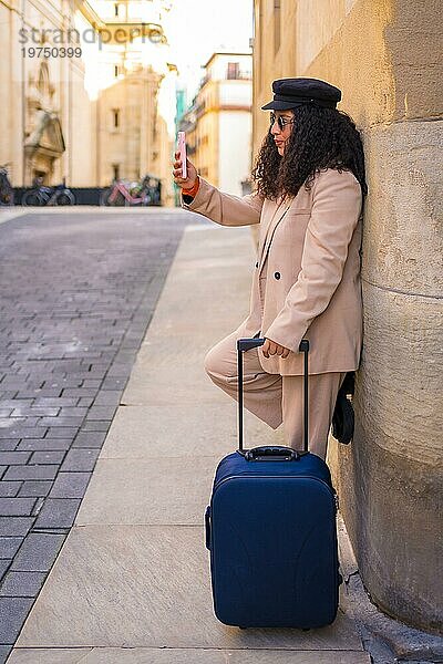 Stilvolle lateinische elegante Frau  die selfie beim Warten mit Gepäck im Freien in einem sonnigen Tag des Winters