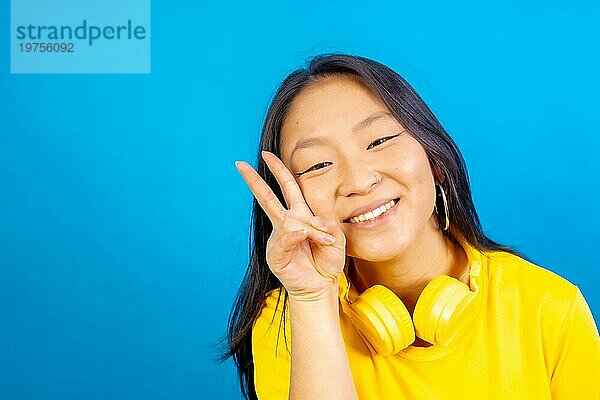 Studiofoto mit blauem Hintergrund einer lächelnden chinesischen Frau mit Kopfhörern gestikulieren Friedenszeichen