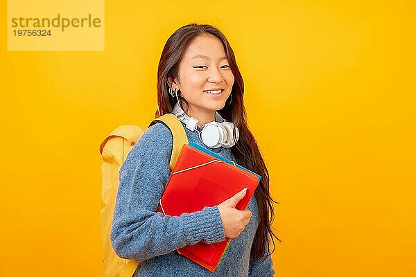 Studiofoto mit gelbem Hintergrund eines chinesischen Studenten mit Kopfhörern  Tasche und Ordnern
